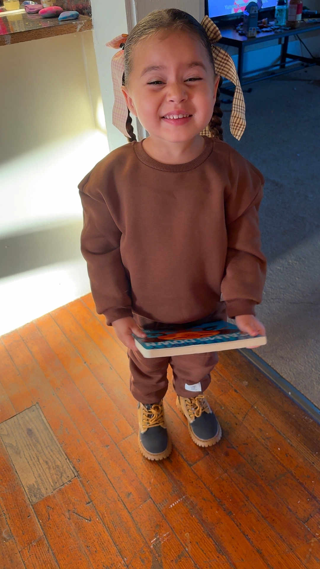 Izabella is registered to the contest to win money with this photo: child, smiling, braids, hair_bow, brown_clothing, boots, book, wooden_floor, indoor, person, happy, toddler, holding, casual_wear, sunlight, flooring, portrait, cute, home, standing