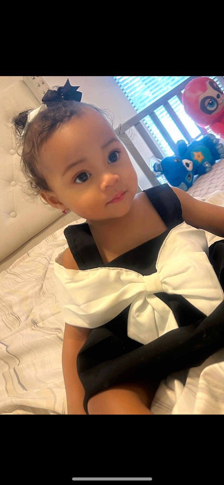 Ascella is registered to the contest to win money with this photo: toddler, child, baby, black_dress, white_bow, hair_bow, bed, plush_toys, stuffed_animals, indoor, portrait, cute, smiling, person, young_child, cozy, window, natural_light, bedroom, decor