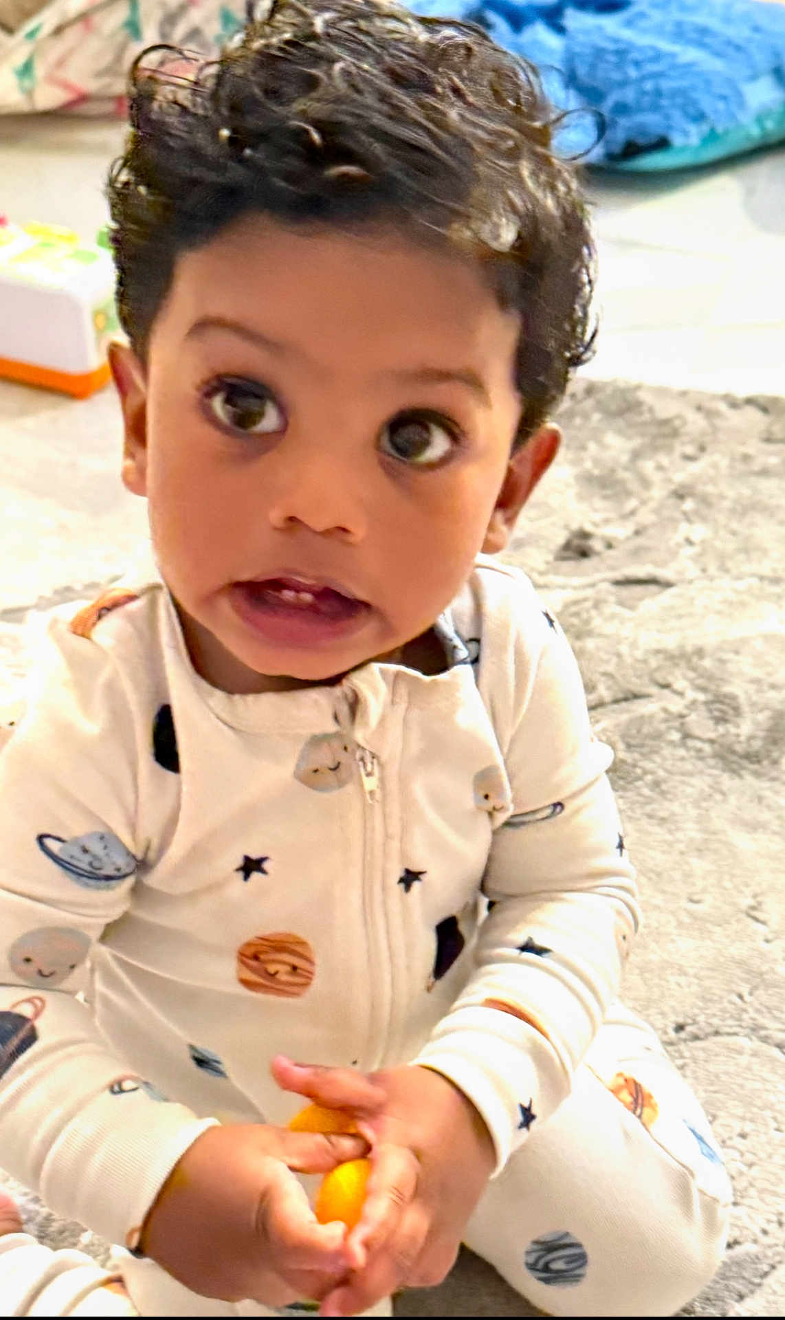 Ethan is registered to the contest to win money with this photo: toddler, child, baby, indoor, carpet, toy, hands, onesie, space_theme, curly_hair, wide_eyes, expression, sitting, cute, casual, cozy, playful, floor, portrait, young_child