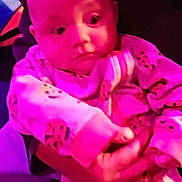 Samantha is registered to the contest to win money with this photo: baby, infant, onesie, hand, pink_light, purple_light, patterned_clothing, child, person, holding, blurred_background, soft_lighting, cute, indoor, sleepwear, small_hands, portrait, newborn, young_child, human