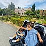 Elham is registered to the contest to win money with this photo: toddler, child, double_stroller, blue_dress, outdoor, sidewalk, greenery, trees, building, sky, cloudy, park, person, footwear, socks, hand, curious, happy, nature, urban