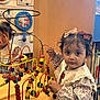 Elham is registered to the contest to win money with this photo: child, toddler, toy, bead_maze, playroom, bookshelf, book, curly_hair, bow, floral_dress, wooden_toy, educational_toy, indoor, play, colorful, person, furniture, face, expression, hands