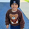 child, toddler, smiling, portrait, outdoor, playground, beanie, hat, sweatshirt, teddy_bear, jeans, boots, standing, full_body, happy, play_area, blue_floor, fence, daylight, fashion