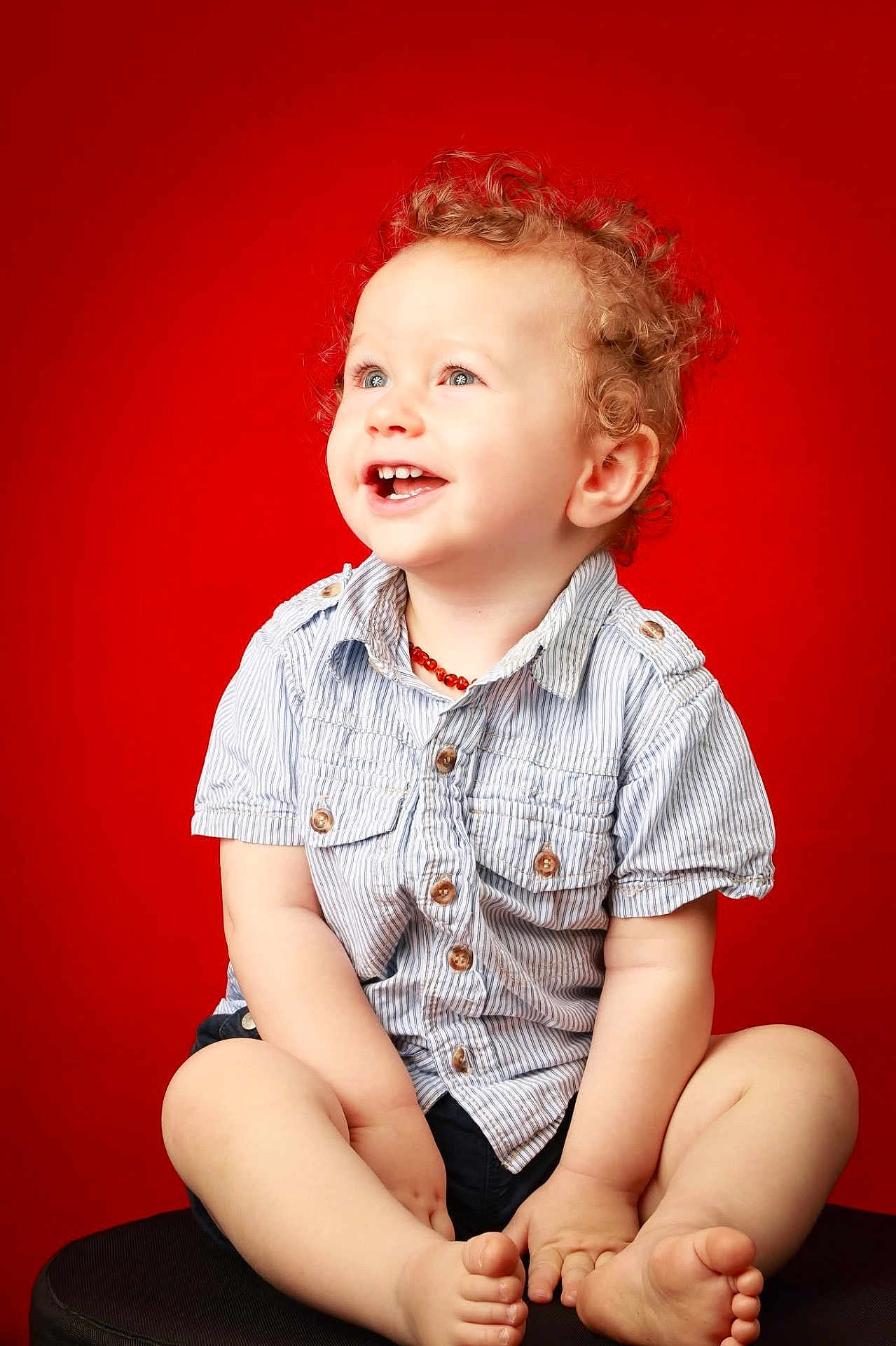 Alan a rejoint le concours — aidez-le/la à gagner de superbes lots ! toddler, child, curly_hair, smiling, blue_eyes, striped_shirt, shorts, barefoot, sitting, red_background, studio_photo, happy, cute, portrait, button_up, young_child, innocent, joyful, indoor, baby