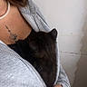 person, hand, grey_hoodie, black_kitten, tattoo, pearl_necklace, indoor, wall, zipper, holding, snuggling, young_adult, casual_clothing, skin, fingers, comfort, pet, animal, cuddle, closeup