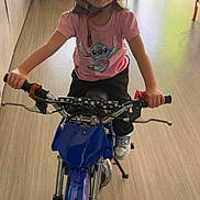 Kenza a rejoint le concours — aidez-le/la à gagner de superbes lots ! bike, black_pants, blue_bike, child, floor, handlebars, helmet, helmet_strap, indoor, person, pink_shirt, room, sliding_door, smile, sneakers, stitch_cartoon, toy, window, wood_floor, young_child