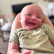 Parker is registered to the contest to win money with this photo: baby, smiling, infant, child, person, face, happy, clothing, patterned_shirt, skin, legs, blurred_background, indoor, cute, portrait, young_child, baby_clothes, comfort, relaxing, closeup