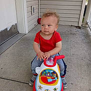 Avery joined the competition — help win amazing prizes! toddler, child, toy_car, red_shirt, sneakers, porch, concrete, curly_hair, sidewalk, house, door, outdoor, play, casual_clothing, young_child, sitting, looking_away, daytime, expression, hands