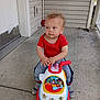 toddler, child, toy_car, red_shirt, sneakers, porch, concrete, curly_hair, sidewalk, house, door, outdoor, play, casual_clothing, young_child, sitting, looking_away, daytime, expression, hands