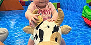 Amarah joined the competition — help win amazing prizes! baby, child, plush_toy, cow, indoor_play_area, blue_floor, tattoo, arm, adult, phone, playground, toy, child_playing, person, colorful, fun, smiling, curious, support, indoor