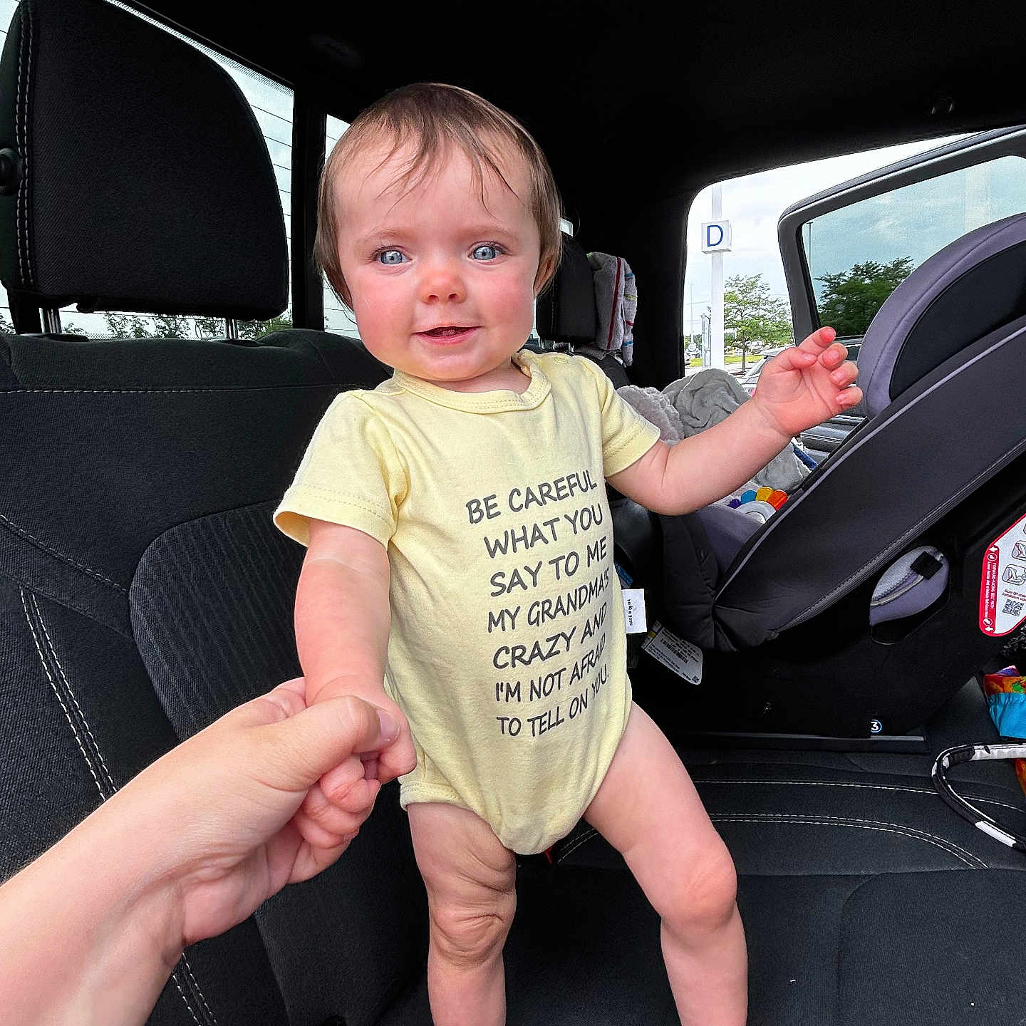 Amarah joined the competition — help win amazing prizes! adult_hand, baby, blue_eyes, car_interior, car_seat, casual, child, clothing, daylight, hand_holding, infant, onesie, parking_lot, person, seat, smiling, standing, toy, vehicle, window