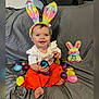 baby, child, bunny_ears, easter_eggs, plush_toy, smiling, orange_pants, white_shirt, gray_blanket, blue_egg, purple_egg, yellow_egg, pink_egg, golden_egg, sitting, indoors, cute, holiday, celebration, happy