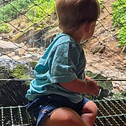 Carson is registered to the contest to win money with this photo: back_view, barefoot, blue_shirt, candid, child, curious, daylight, greenery, metal_grate, nature, outdoor, raindrops, rocks, scenic, shorts, sitting, toddler, water, waterfall, wire_mesh