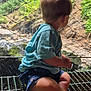 back_view, barefoot, blue_shirt, candid, child, curious, daylight, greenery, metal_grate, nature, outdoor, raindrops, rocks, scenic, shorts, sitting, toddler, water, waterfall, wire_mesh