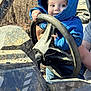 child, toddler, hoodie, blue_clothing, steering_wheel, muddy, outdoor, forest, person, baby, hand, vehicle, adult_arm, nature, playful, curious, portrait, daylight, expression, casual