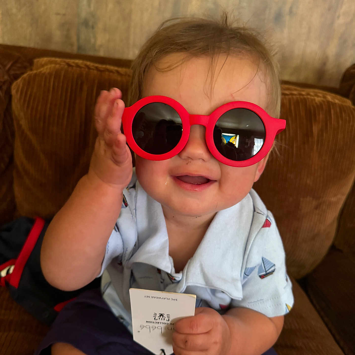 Carson is registered to the contest to win money with this photo: baby, child, clothing, couch, cute, drool, face, hand, happy, indoor, person, playful, red, seat, shirt, shorts, smiling, sunglasses, toddler, wood_wall