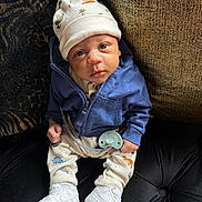 Micah is registered to the contest to win money with this photo: baby, infant, hat, blue_jacket, pacifier, white_shoes, couch, black_couch, patterned_pants, cute, child, sitting, indoors, portrait, face, clothing, footwear, expression, cozy, small
