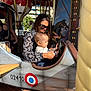 airplane_ride, amusement_ride, carousel, child, clothing, daylight, family, fun, horse_figure, hot_air_balloon, outdoor, parent, play, ride, smile, sunglasses, toddler, toy, woman, wooden_floor