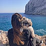 person, dog, rocky_cliff, ocean, sea, sky, blue_sky, long_hair, casual_clothing, sitting, outdoor, nature, pet, fluffy_dog, scruffy_dog, relaxing, sunlight, footwear, calm, scenery