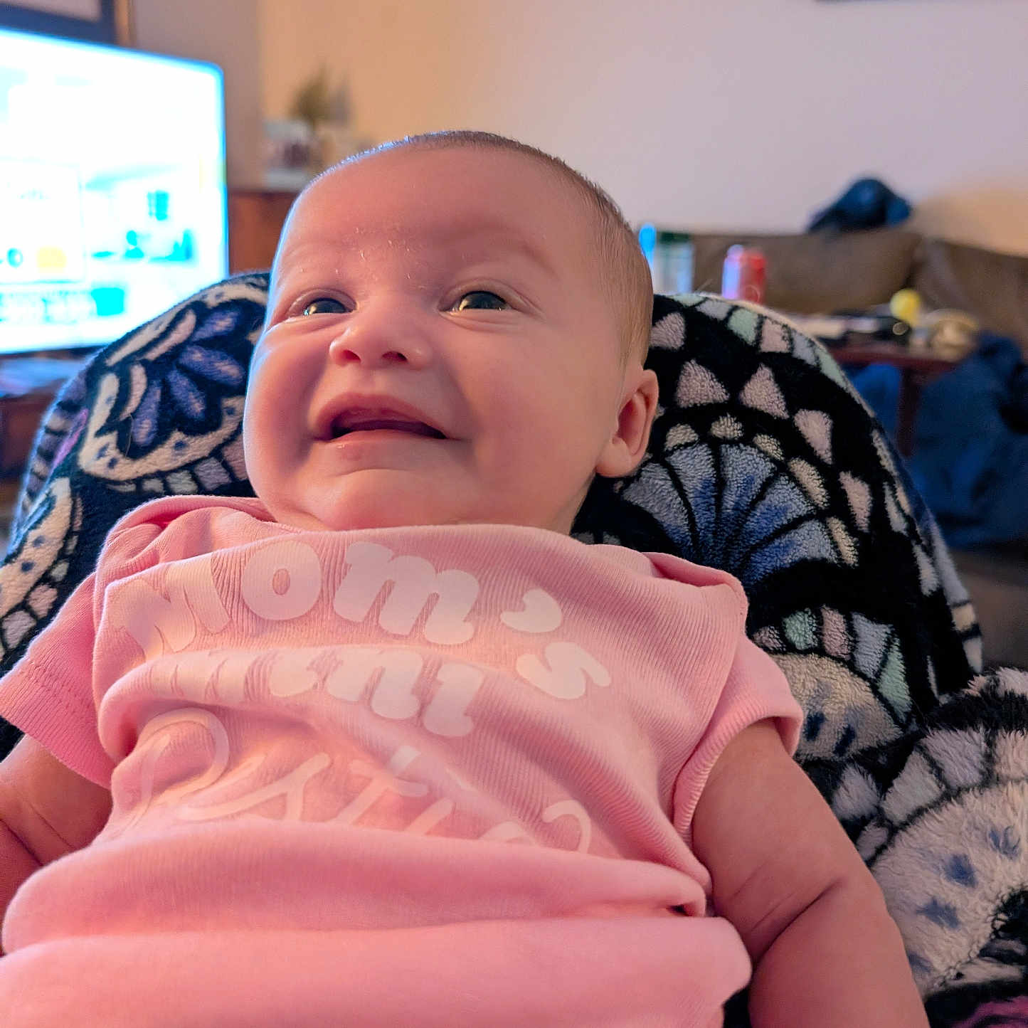 Mackenzie is registered to the contest to win money with this photo: baby, infant, smiling, pink_clothing, onesie, blanket, face, portrait, indoor, living_room, person, happy, cute, child, young, head, skin, expression, seated, cozy