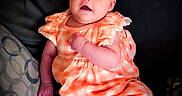 Mackenzie joined the competition — help win amazing prizes! baby, infant, child, orange_clothing, headband, bow, couch, pillow, patterned_pillow, smiling, cute, indoors, portrait, person, sitting, plaid, comfortable, bright_eyes, soft_lighting, relaxed