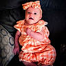 baby, infant, child, orange_clothing, headband, bow, couch, pillow, patterned_pillow, smiling, cute, indoors, portrait, person, sitting, plaid, comfortable, bright_eyes, soft_lighting, relaxed