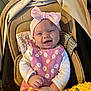 baby, smiling, pink_bow, pink_dress, floral_pattern, stroller, blanket, knitted_blanket, yellow_blanket, cushion, infant, happy, child, headband, clothing, portrait, indoors, cute, baby_girl, seated