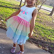 Ava is registered to the contest to win money with this photo: child, girl, smile, outdoor, dress, unicorn, bow, curly_hair, tutu, pastel_colors, footwear, sidewalk, grass, tree, sunlight, happy, standing, nature, casual, portrait