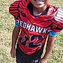 child, boy, football_uniform, red_uniform, sports, outdoor, grass, smiling, youth, athlete, team, jersey, cleats, socks, black_shoes, field, sunlight, fence, person, game