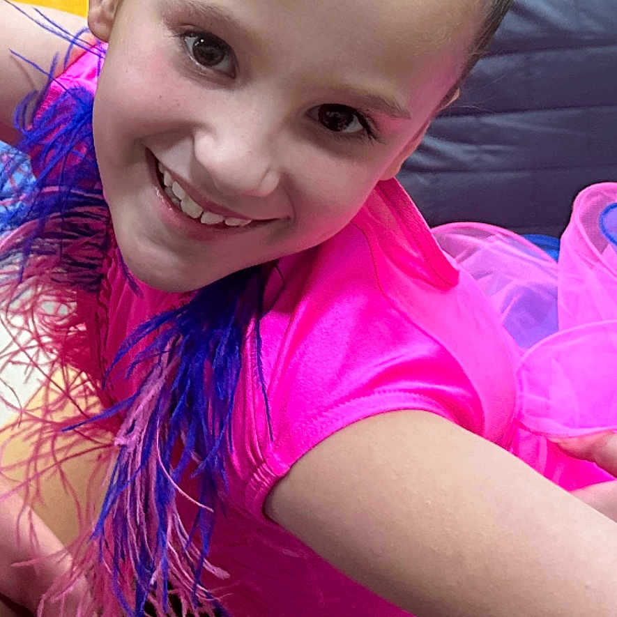 Aleigha is registered to the contest to win money with this photo: bench, cheerful, child, closeup, costume, dance, feathers, floor, girl, gymnasium, hair_up, happy, performance, person, pink_clothing, portrait, posing, purple_feathers, smiling, young