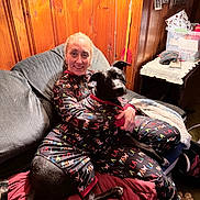 Aries is registered to the contest to win money with this photo: animal, black_dog, blanket, couch, cozy, dog, furniture, home, indoor, lamp, living_room, matching_outfits, pajamas, person, pet, relaxed, seated, smiling, woman, wood_paneling