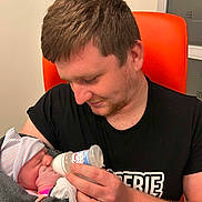 Tiago participe au concours pour gagner de l'argent avec cette photo : baby, newborn, father, feeding, bottle, hat, blanket, indoor, person, man, child, holding, orange_chair, cozy, love, family, care, gentle, portrait, tender_moment