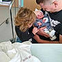newborn, baby, toddler, man, hospital, bed, blanket, clothing, holding, family, parent, child, curly_hair, smiling, indoor, floor, wheelchair, caring, gentle, tender_moment