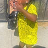 child, toddler, standing, outdoor, sunlight, yellow_outfit, cartoon_pattern, braids, earrings, face_paint, sparkle, smiling, hands, finger_tasting, purse, sandal, sidewalk, patio, shadow, portrait