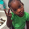 child, toddler, portrait, smiling, pouting, peace_sign, bathroom, indoor, green_shirt, shorts, earrings, braided_hair, dark_skin, bathtub, countertop, bath_products, bath_stones, toy_stroller, standing, playful
