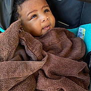 Princess is registered to the contest to win money with this photo: baby, child, towel, brown, curly_hair, stroller, blanket, face, portrait, infant, cute, wrapped, cozy, looking_up, indoors, soft_texture, person, young, comfortable, expression
