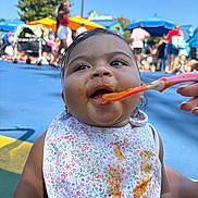 Araiya Lynn is registered to the contest to win money with this photo: baby, child, feeding, bib, food, messy, spoon, outdoor, people, crowd, summer, daylight, colorful, umbrella, happy, open_mouth, clothing, skin, portrait, closeup