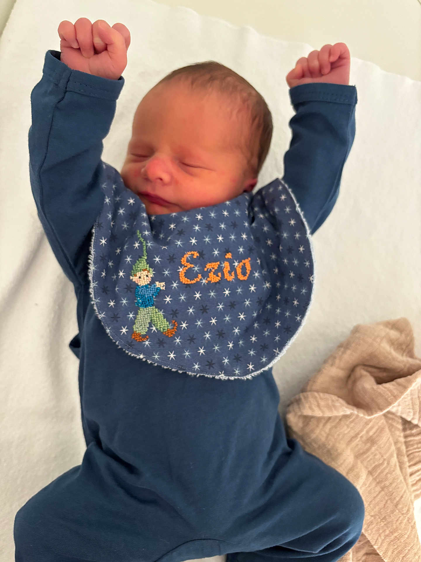 Ezio participe au concours pour gagner de l'argent avec cette photo : newborn, baby, infant, sleeping, blue_clothing, bib, embroidered, name, arms_up, soft_blanket, white_background, peaceful, cute, lying_down, fabric, closeup, child, person, indoors, relaxing