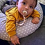 baby, pacifier, yellow_sweater, polka_dot, cushion, indoor, carpet, hand, face, child, curious, laying_down, clothing, comfort, young_child, person, soft_furniture, home, baby_accessory, portrait