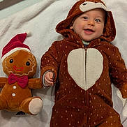 éliot a rejoint le concours — aidez-le/la à gagner de superbes lots ! baby, child, plush_toy, gingerbread, santa_hat, red_bow_tie, onesie, brown_onesie, heart_pattern, smiling, lying_down, white_blanket, soft_texture, cute, holiday, christmas, festive, hand_holding, indoors, portrait