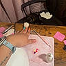 cat, black_cat, white_paw, chair, wooden_table, hand, colorful_nails, nail_art_tools, pink_mat, curtain, indoor, pet, playful, paw, person, watch, tattoo, bottle, pen, relaxed