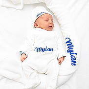 Mylan a rejoint le concours — aidez-le/la à gagner de superbes lots ! baby, newborn, sleeping, white_clothing, hat, blanket, soft_texture, blue_text, name_embroidery, cozy, infant, peaceful, portrait, lying_down, cute, child, comfort, soft_lighting, studio_photo, white_background