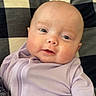 baby, infant, face, smile, blue_eyes, clothing, purple_outfit, chubby_cheeks, close_up, blanket, pattern, head, skin, person, cute, happy, young_child, portrait, indoor, soft_light