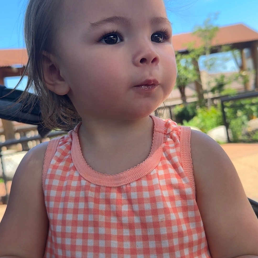 Kyla joined the competition — help win amazing prizes! blue_sky, casual, checkered, child, curious, daytime, dress, expression, face, hair, hand, nature, outdoor, peach, person, portrait, snack, sunlight, toddler, young_child