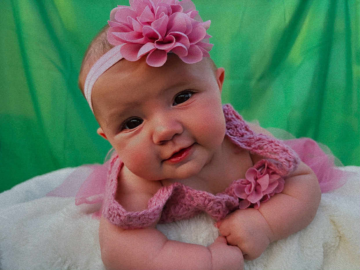 Madison is registered to the contest to win money with this photo: baby, infant, pink, headband, flower, knitted_clothing, blanket, green_background, cute, portrait, smiling, chubby_cheeks, toddler, soft_texture, curious_expression, closeup, child, young, indoors, hands