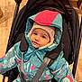 child, toddler, stroller, winter_jacket, hat, indoor, baby, seatbelt, cute, warm_clothing, face, baby_carrier, curious, colorful_clothing, infant, sitting, padded_jacket, young_child, smiling, portrait