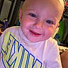 baby, blond_hair, blue_eyes, child, close_up, clothing, drool, face, graphic_t_shirt, happy, home, indoor, infant, photography, playful, portrait, rosy_cheeks, sitting, smiling, warm_lighting