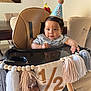 baby, high_chair, party_hat, half_birthday, tassels, banner, indoor, dining_room, table, chair, foot, curly_hair, child, celebration, smiling, cute, wooden_floor, decorations, face, person
