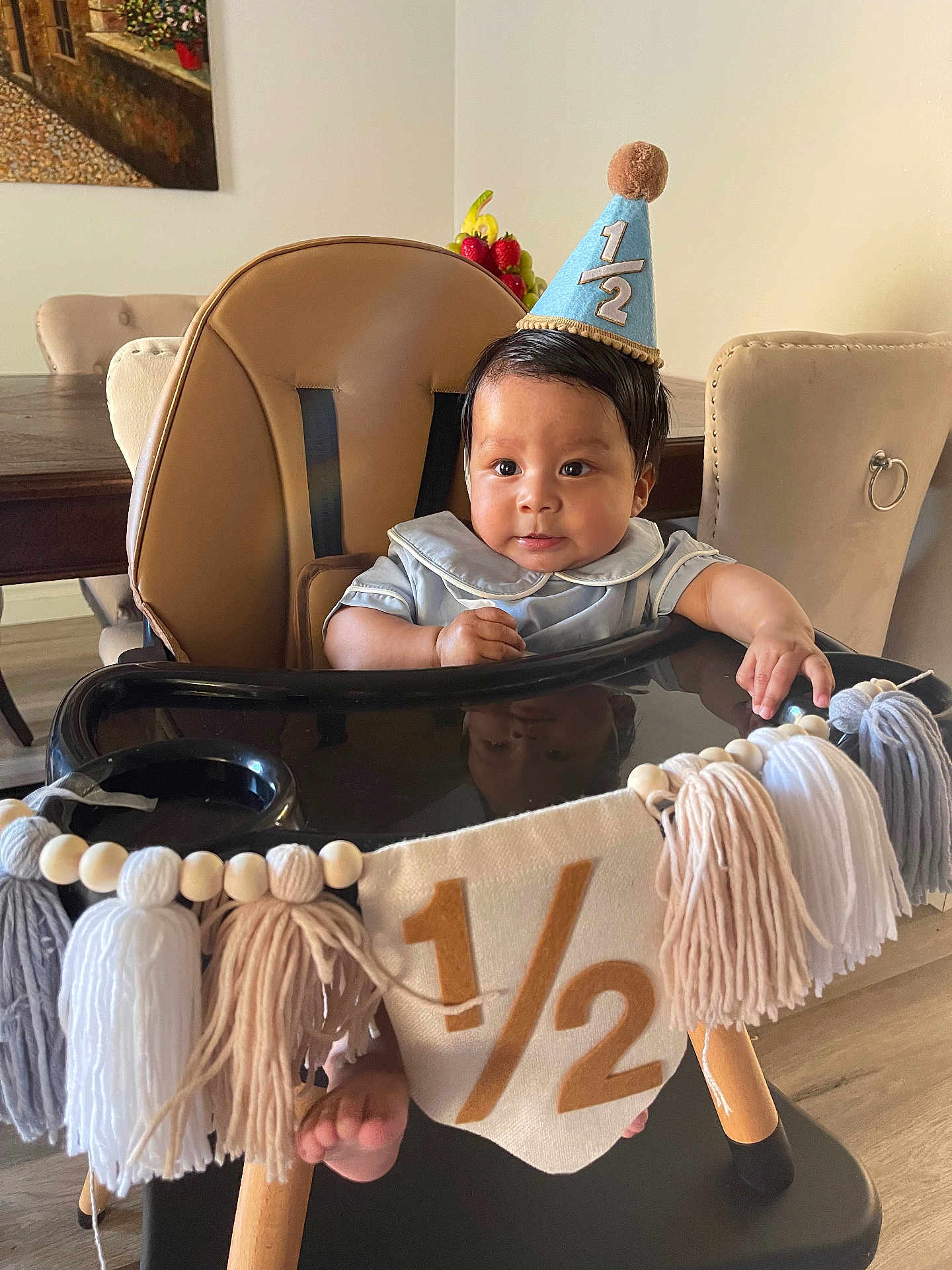 Ryan is registered to the contest to win money with this photo: baby, high_chair, party_hat, half_birthday, tassels, banner, indoor, dining_room, table, chair, foot, curly_hair, child, celebration, smiling, cute, wooden_floor, decorations, face, person