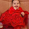 baby, child, smiling, happy, portrait, red_dress, red_clothing, infant, face, eyes, hair, hand, sitting, chair, cushion, blanket, indoor, patterned_fabric, fashion, family_photo